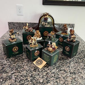 May include: A collection of decorative bear figurines, each posed differently and atop individual green boxes. The bears are various colours, with detailed clothing and accessories. A clock and framed artwork are in the background, with a granite countertop.