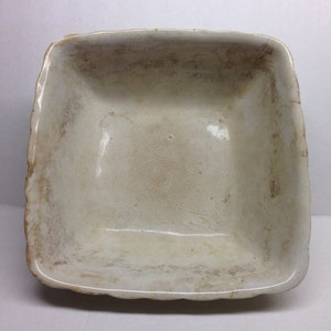 May include: A white ceramic square baking dish with a slightly scalloped edge. The dish has a worn, aged appearance with some brown staining.