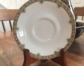 May include: A white saucer with a decorative floral border featuring pink roses and green foliage. The saucer is displayed on a small wooden stand, showcasing its design. The saucer is a vintage piece.