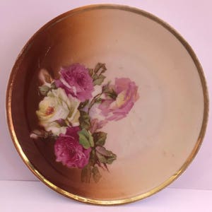May include: A white porcelain plate with a gold rim and a floral design featuring pink and white roses. The plate has a brown gradient background.