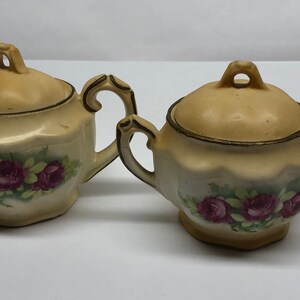 May include: A vintage cream-colored miniature teapot and sugar bowl set. The set features a floral design with burgundy roses and green leaves. Both pieces have a gold trim and ornate handles, perfect for a tea party.
