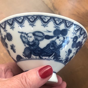 May include: A white porcelain bowl with a blue and white design. The bowl features two figures in traditional Chinese clothing, holding a fan and a stick. The bowl has a scalloped rim and a small foot.