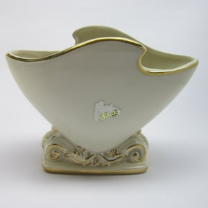 May include: Cream-colored ceramic vase with a unique, asymmetrical shape and gold trim. The base features ornate gold scrollwork. A sticker on the side reads "CHINA". This decorative vase is suitable for displaying flowers or as a standalone accent piece.