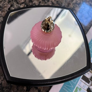 May include: A pink, frosted glass perfume bottle with a gold-colored top sits on a mirrored surface within a black-framed square. The bottle's design features a ribbed texture, and its reflection is visible on the mirror.