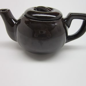 Pode incluir: Bule pequeno de cerâmica castanho escuro com corpo arredondado, bico curvo e pega quadrada. A tampa tem um pequeno botão redondo. O bule é brilhante e está sobre uma superfície branca. Adequado para servir chá.