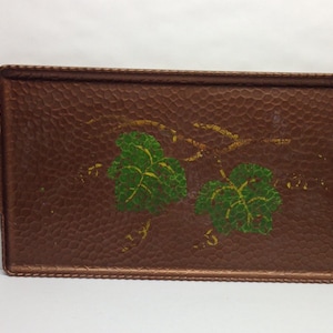 Puede incluir: Una bandeja rectangular marrón con una superficie texturizada y bordes cosidos. La bandeja presenta un diseño pintado de hojas verdes y vides doradas. Tiene dos asas adjuntas.