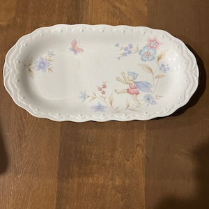 Könnte beinhalten: Weiße ovale Servierplatte mit einem Blumendesign, das eine Fee mit Flügeln, Schmetterlingen und Blumen in Blautönen, Rosa und Grün zeigt.