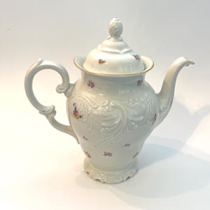 May include: A white porcelain teapot with a lid, handle, and spout. The teapot is decorated with embossed floral patterns and small, colorful flower clusters. The rim of the lid has a gold trim.