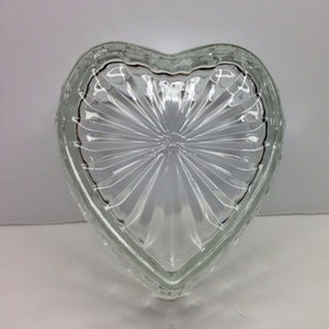 Puede incluir: Jarra de baratijas en forma de corazón de vidrio transparente. El plato tiene un diseño texturizado y acanalado que irradia desde el centro. El borde está delineado con un borde oscuro. El plato está vacío y se encuentra sobre una superficie blanca.