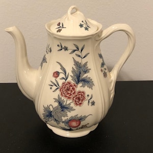 May include: A white ceramic teapot with a floral design in shades of pink, blue, and green. The teapot has a lid and a handle.