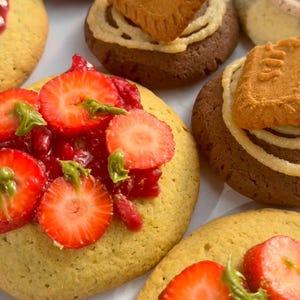 Könnte beinhalten: Nahaufnahme von mehreren dekorierten Keksen. Einige Kekse sind mit geschnittenen Erdbeeren und roter Marmelade belegt, andere mit einem Keks und einer Sahnespirale. Die Kekse liegen auf einer weißen Oberfläche.