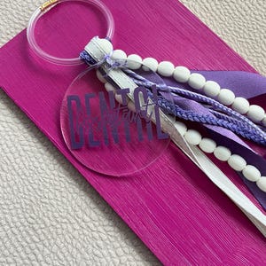Op de afbeelding: Een ronde sleutelhanger van helder acryl met de paarse tekst Dental Assistant. De sleutelhanger is bevestigd aan een doorzichtige kunststof ring en bevat een decoratieve kwast van witte houten kralen, paars lint en gevlochten paars koord.