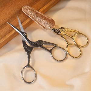 Könnte beinhalten: Zwei Paar dekorative Stickschere. Ein Paar ist silberfarben mit dem Namen "Daisy" auf der Klinge eingraviert. Das andere Paar ist goldfarben. Eine goldglitzernde Schutzhülle ist im Lieferumfang enthalten.