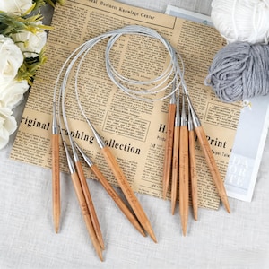 May include: A collection of wooden circular knitting needles with metal tips and flexible cords. The needles are arranged on a newspaper background, with balls of white and gray yarn visible. Some needles have engraved text.