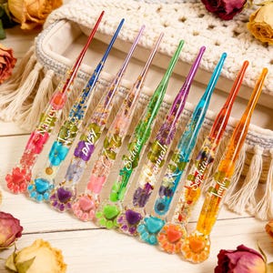 May include: A collection of eight crochet hooks with clear resin handles, each containing dried flowers and a name. The hooks have colorful tips and paw-shaped bases with heart-shaped accents. The names include Nancy, Daisy, and Sophia.