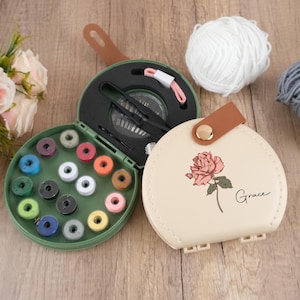 May include: A cream-coloured sewing kit with a rose graphic and the word "Grace." The kit is open, revealing spools of thread in various colours, needles, and other sewing tools. A ball of white yarn is also visible.