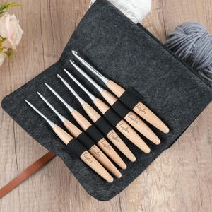 May include: A collection of crochet hooks with wooden handles and silver metal tips, presented in a grey felt case. Each handle is engraved with the name "Taylor". The case is open, showing the hooks and a brown leather strap.