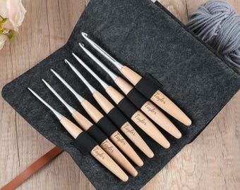Può includere: Un set di uncinetti con manici in legno e punte in metallo argentato, disposti in un astuccio di feltro grigio. Ogni manico è inciso con il nome "Taylor". L'astuccio è aperto, rivelando gli uncinetti e un cinturino in pelle marrone.