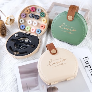 May include: A beige sewing kit with a variety of colorful thread spools, needles, and scissors. Two round cases, one green and one beige, are also visible, each with the words "THINGS WITH love" printed on them. The green case has a brown leather strap.