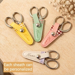 May include: A selection of small scissors with silver blades and colourful leather sheaths. The sheaths are in yellow, green, pink, and white, each with the word "Name" embossed. The text "Each sheath can be personalised" is also visible.