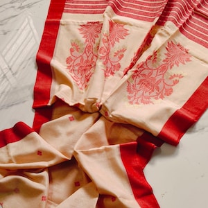 Op de afbeelding: Een crèmekleurig textiel met een opvallende rode rand en rode streeppatronen. De stof is voorzien van ingewikkelde rode bloemmotieven die in het materiaal zijn geweven, met kleine rode decoratieve accenten verspreid over het onderste gedeelte.