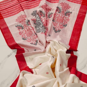 Op de afbeelding: Een crèmekleurig textiel met een brede rode rand en horizontale rode strepen. De stof is versierd met grote, ingewikkelde bloemmotieven in tinten rood en donkergrijs, samen met kleine, verspreide donkergrijze en rode bloemaccenten.