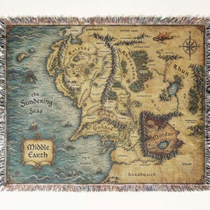 May include: A woven throw blanket featuring a detailed map of Middle Earth, including the Sundering Seas, Rohan, Gondor, and Mordor. The design includes a compass rose, a dragon illustration, and text labels for various regions, finished with a fringed edge.
