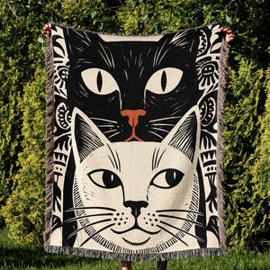 May include: A black and cream-colored throw blanket featuring two stylized cat faces. The top cat is black with white eyes and a brown nose, while the bottom cat is cream-colored with black details. The blanket has a fringed edge.