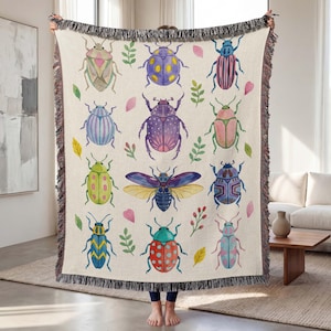 May include: Beige throw blanket featuring a repeating pattern of colorful watercolor beetles, leaves, and floral accents. The blanket has a fringed edge and is held up by a person.
