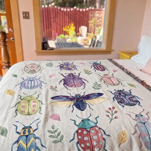 Puede incluir: Manta beige con ilustraciones de acuarela de varios escarabajos en tonos azules, verdes, rosas y morados. La manta tiene un borde con flecos y est&aacute; colocada sobre una cama. El fondo incluye una ventana y un vistazo al exterior.