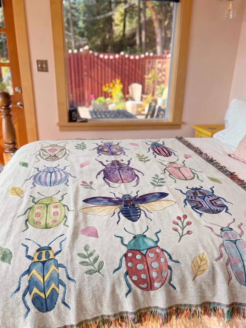 Puede incluir: Manta beige con ilustraciones de acuarela de varios escarabajos en tonos azules, verdes, rosas y morados. La manta tiene un borde con flecos y est&aacute; colocada sobre una cama. El fondo incluye una ventana y un vistazo al exterior.