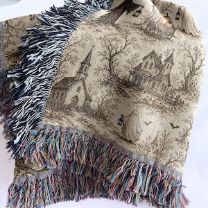May include: A woven throw blanket featuring a beige background with a dark brown print of a haunted scene including a church, a house, bare trees, a ghost holding a lantern, and bats. The blanket has a decorative fringe edge with multi-colored threads.