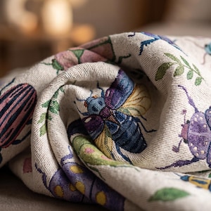 Puede incluir: Tela beige con un patr&oacute;n repetido de escarabajos coloridos en tonos de azul, morado, rosa y verde. La tela est&aacute; doblada, mostrando ilustraciones detalladas de insectos y un tejido texturizado.