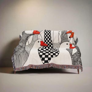 May include: A cream-colored throw blanket draped over a sofa, featuring a whimsical design of geese with red beaks. The blanket has a black and white checkered pattern and fringe along the bottom edge. The geese are outlined in black.