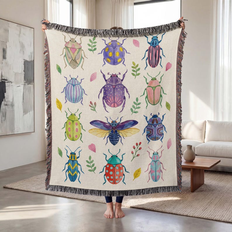 May include: Beige throw blanket featuring a repeating pattern of colorful watercolor beetles, leaves, and floral accents. The blanket has a fringed edge and is held up by a person.