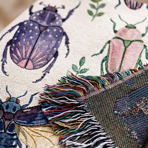 Puede incluir: Manta beige con un patr&oacute;n repetido de escarabajos coloridos y follaje verde. La manta tiene un borde con flecos multicolores. Los escarabajos son morados, rosas y azules.