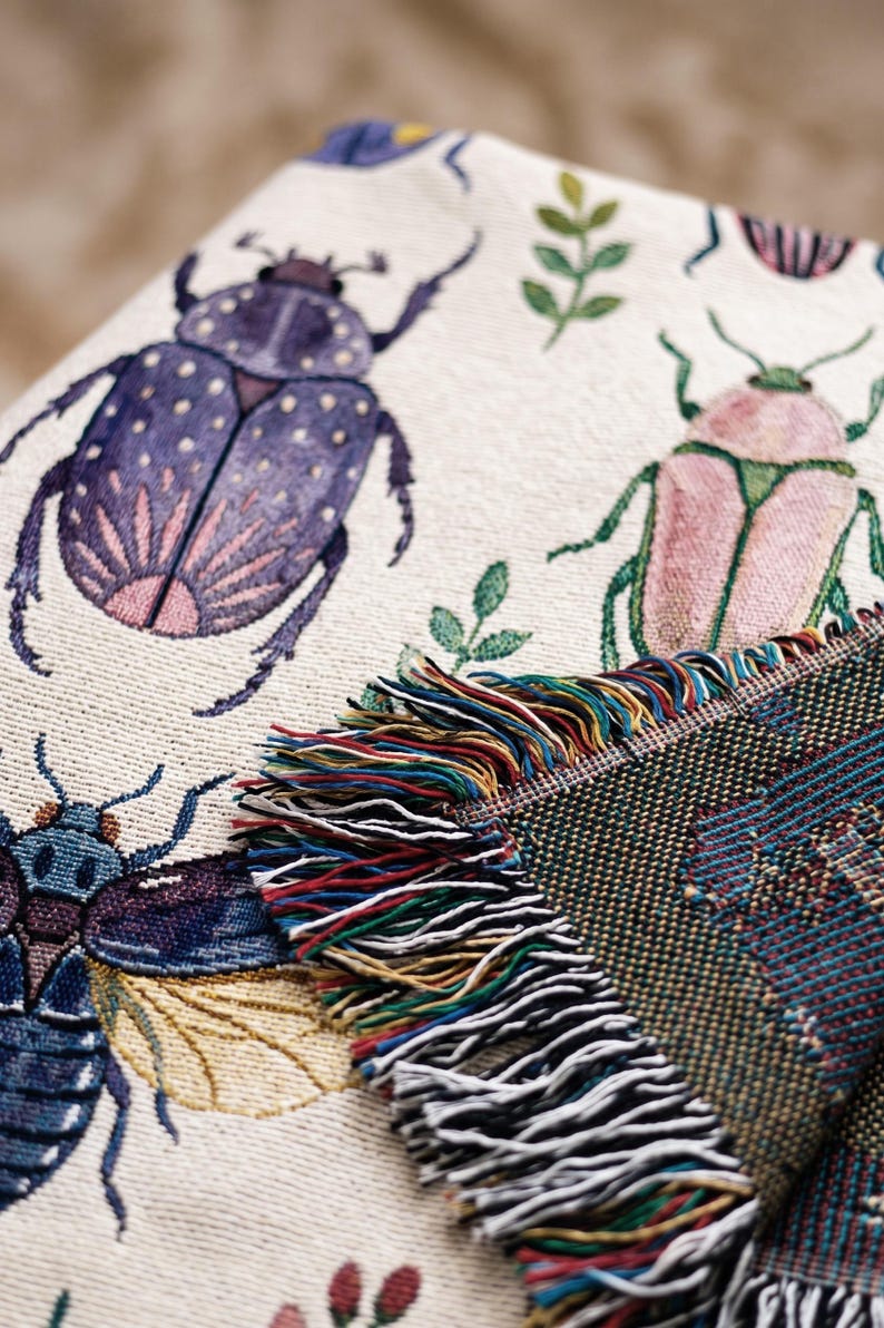 Puede incluir: Manta beige con un patr&oacute;n repetido de escarabajos coloridos y follaje verde. La manta tiene un borde con flecos multicolores. Los escarabajos son morados, rosas y azules.