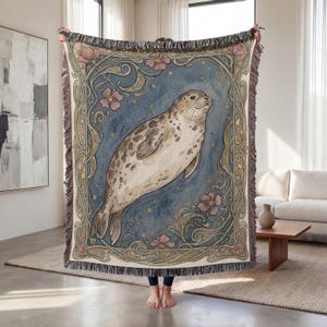 May include: A woven throw blanket with a seal illustration. The seal is in a blue and beige colour scheme, framed by floral and scrollwork designs. The blanket has a fringed edge. The blanket is held up by a person.