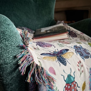 Puede incluir: Manta beige con ilustraciones de insectos coloridos, incluyendo escarabajos y mariposas, sobre un sill&oacute;n verde. La manta tiene un borde con flecos de hilos multicolores. Un libro descansa sobre la manta.