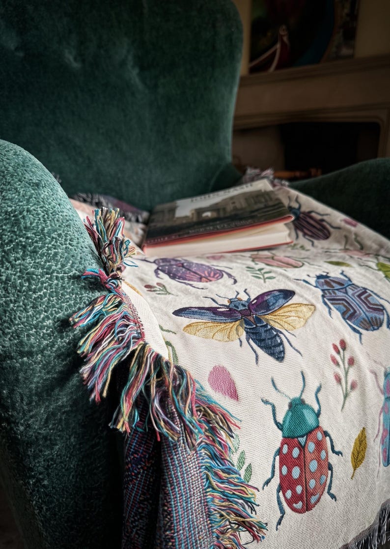 Puede incluir: Manta beige con ilustraciones de insectos coloridos, incluyendo escarabajos y mariposas, sobre un sill&oacute;n verde. La manta tiene un borde con flecos de hilos multicolores. Un libro descansa sobre la manta.
