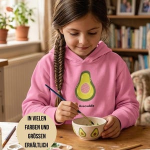 Könnte beinhalten: Ein junger Mensch trägt einen rosa Kapuzenpullover mit einem Avocado-Motiv und dem Wort "Avocuddle". Die Person bemalt eine kleine Schüssel mit einem Avocado-Design. Eine Farbpalette und Pinsel liegen auf dem Tisch. Der Text "IN VIELEN FARBEN UND GRÖSSEN ERHÄLTLICH" befindet sich in einem Kreis.