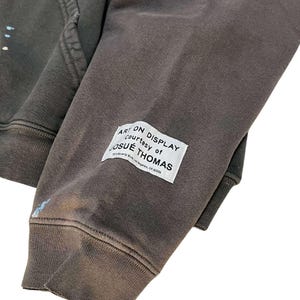 May include: Close-up of a dark gray hoodie sleeve with a white rectangular label. The label reads "ART ON DISPLAY courtesy of JOSUE THOMAS" with an address below. The fabric has a distressed look with paint splatters.