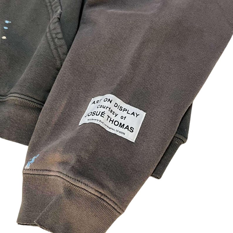 May include: Close-up of a dark gray hoodie sleeve with a white rectangular label. The label reads "ART ON DISPLAY courtesy of JOSUE THOMAS" with an address below. The fabric has a distressed look with paint splatters.