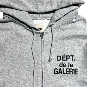 May include: A gray zip-up hoodie with a hood and drawstring. The hoodie features the text "D&Eacute;PT. de la GALERIE" in black block letters on the front. The zipper is silver.