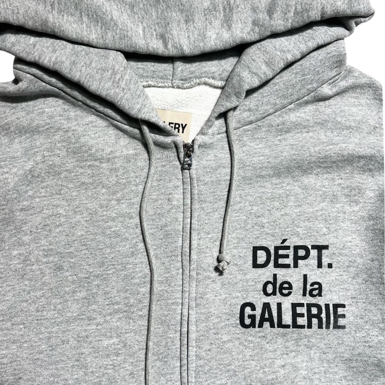 May include: A gray zip-up hoodie with a hood and drawstring. The hoodie features the text "D&Eacute;PT. de la GALERIE" in black block letters on the front. The zipper is silver.