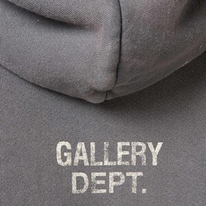 May include: Close-up of a gray hoodie with the text "GALLERY DEPT." printed in white. The hoodie's hood is visible at the top of the frame, with the text centered below. The fabric appears to be a soft, textured material.