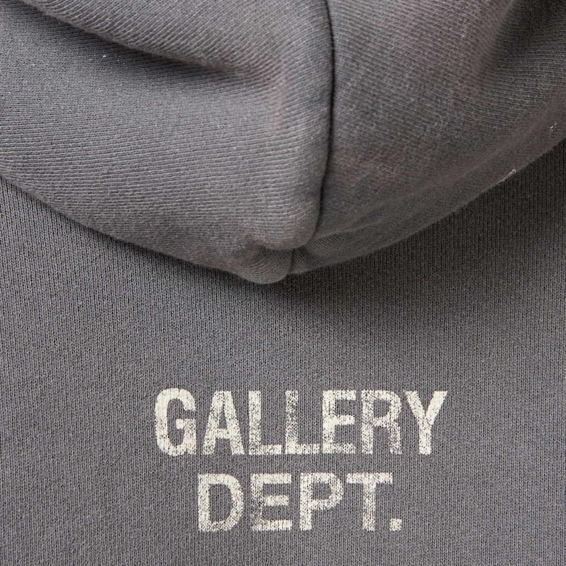 May include: Close-up of a gray hoodie with the text "GALLERY DEPT." printed in white. The hoodie's hood is visible at the top of the frame, with the text centered below. The fabric appears to be a soft, textured material.