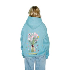 Puede incluir: Sudadera con capucha azul claro con un ramo de flores bordado en la espalda. El ramo presenta flores rosas, blancas y amarillas con hojas verdes. El texto "GIFT GIVING" está debajo.