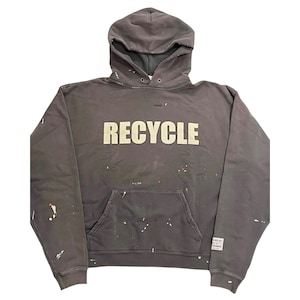 May include: A dark gray hoodie with the word "RECYCLE" in large, off-white block letters across the chest. The hoodie has a front pocket and a drawstring hood. Paint splatters in various colors are visible on the fabric.
