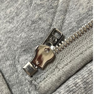 May include: Close-up of a silver zipper on a gray fabric. The zipper pull features a logo. The fabric has a textured appearance. The zipper teeth are visible and metallic.